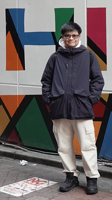 A young man with glasses, wearing a dark hooded jacket over a white hoodie, beige cargo pants, and black boots, stands with his hands in his pockets in front of a colorful geometric mural. He smiles at the camera, exuding a relaxed and approachable demeanor. The pavement beneath him has a painted "No Smoking" sign.